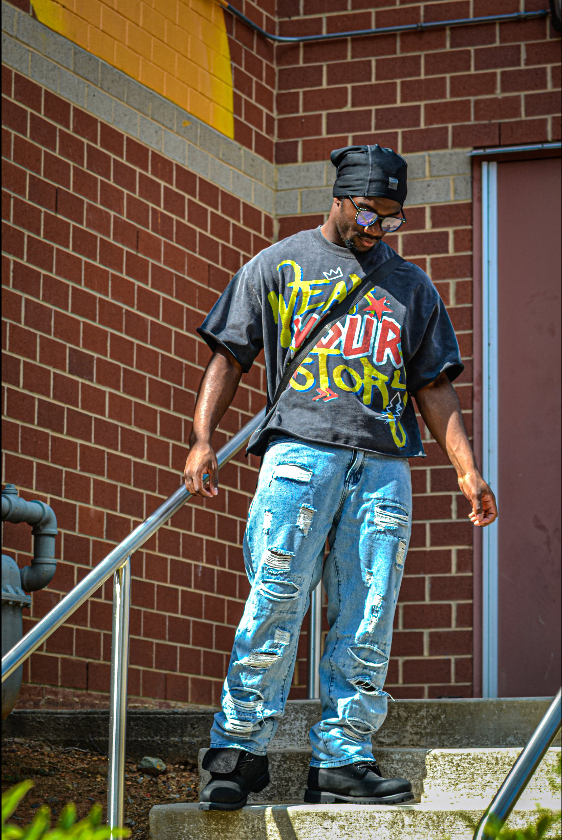 Man wearing a graphic t-shirt and ripped jeans standing on steps against a brick wall.